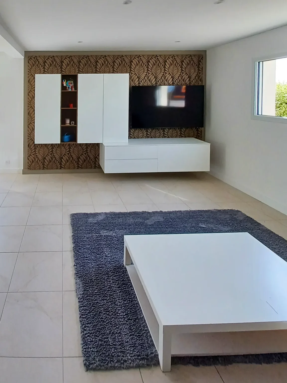 Salon rénové avec table basse blanche, tapis gris et meuble TV suspendu sur papier peint feuillage